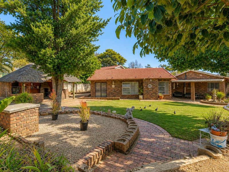 A well-maintained rear garden featuring a brick-faced house, a traditional thatched lapa, and established landscaping with paved pathways.