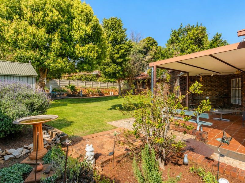 A spacious rear garden featuring a covered brick-paved patio, established trees, lawn area, and a garden shed, providing a private outdoor living space.