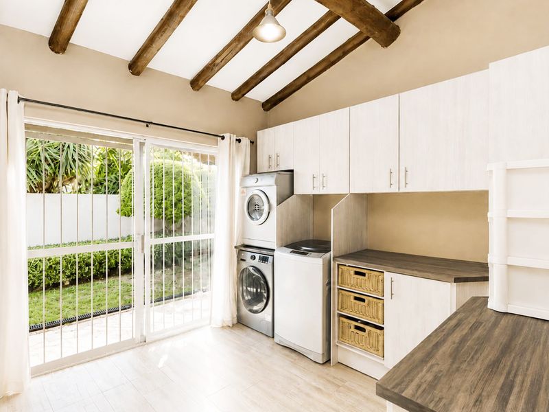 A bright and functional laundry room featuring built-in cabinetry, laminate countertops, exposed timber ceiling beams, and direct access to the garden via sliding doors.