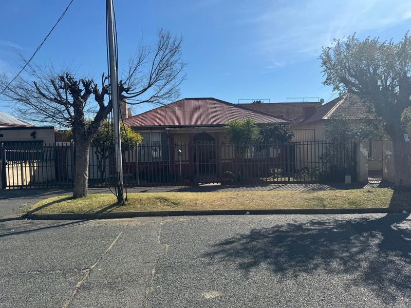 Front elevation of a single-story house featuring a corrugated metal roof, brick facade, and secure metal perimeter fencing.