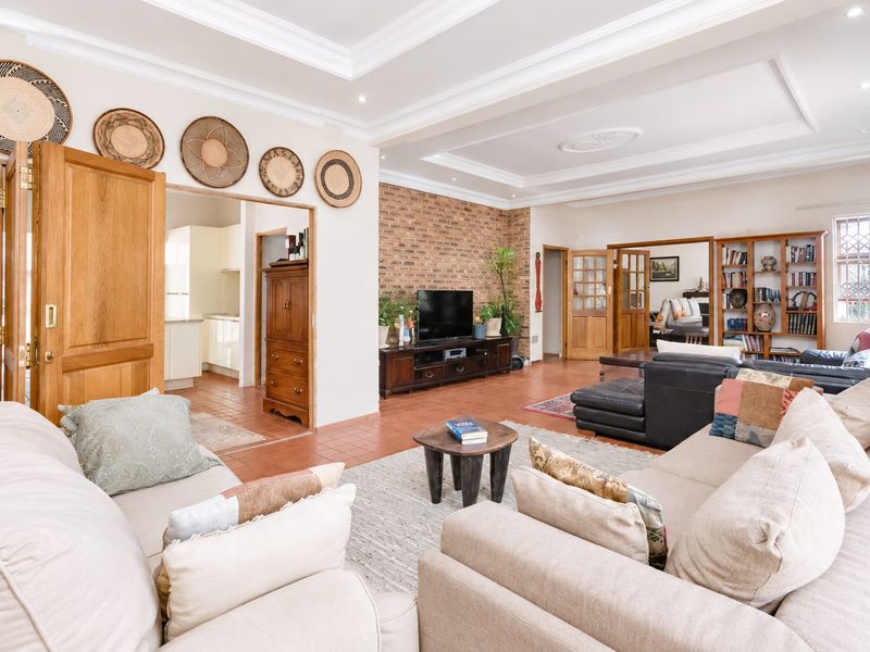 Spacious open-plan living area featuring a distinctive exposed brick wall, recessed tray ceilings, and terracotta flooring, connecting seamlessly to the kitchen and adjacent rooms.