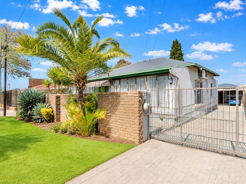 Front view of a residential property featuring a paved driveway, metal security gate, brick boundary wall, and a corrugated metal roof.
