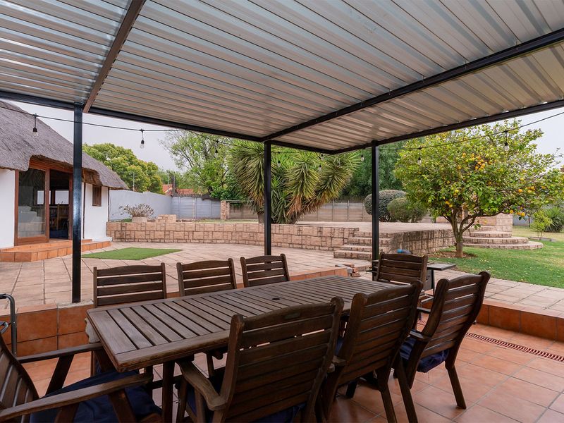 A covered outdoor patio area featuring tiled flooring, a corrugated metal roof, and direct access to a landscaped garden with a brick retaining wall.
