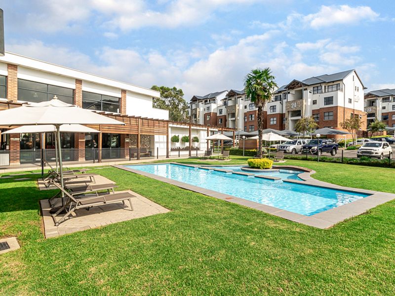 A well-maintained communal swimming pool area featuring a paved deck, landscaped lawn, and timber pergola structure within a modern apartment complex.