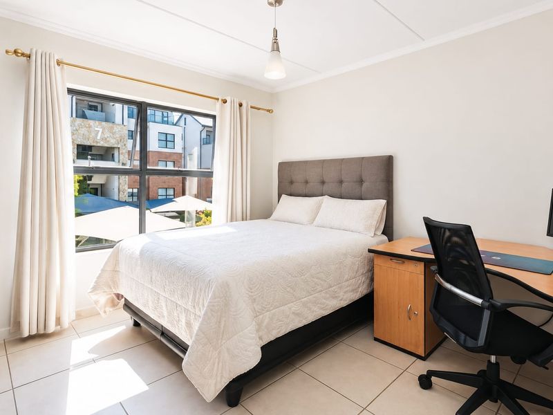 A bright, well-maintained bedroom featuring tiled flooring, neutral walls, and a large window providing natural light and views of the complex.