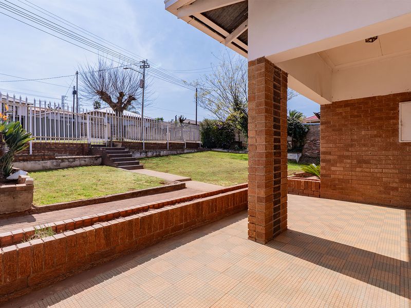 A covered patio area featuring brick pillars and tiled flooring, overlooking a tiered garden with brick retaining walls and perimeter fencing.