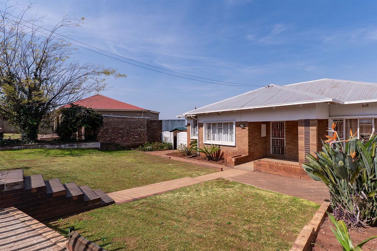 Rear view of a brick house featuring a lawn, concrete pathways, brick steps, and a corrugated metal roof.
