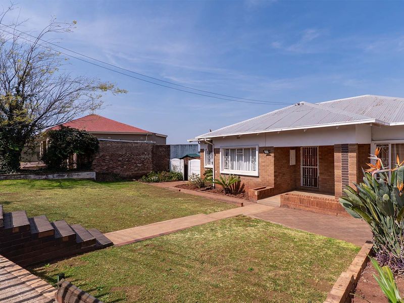 Rear view of a brick house featuring a lawn, concrete pathways, brick steps, and a corrugated metal roof.