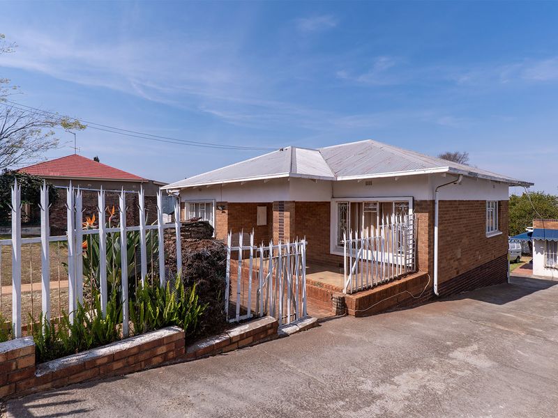 A single-story brick house featuring a corrugated metal roof, a covered front porch, and secure metal perimeter fencing with a concrete driveway.