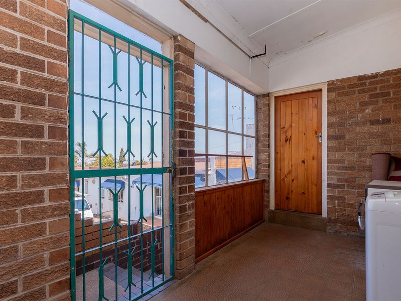An enclosed balcony or sunroom featuring exposed brick walls, large windows, and a security gate, offering a functional space with potential for modernization.
