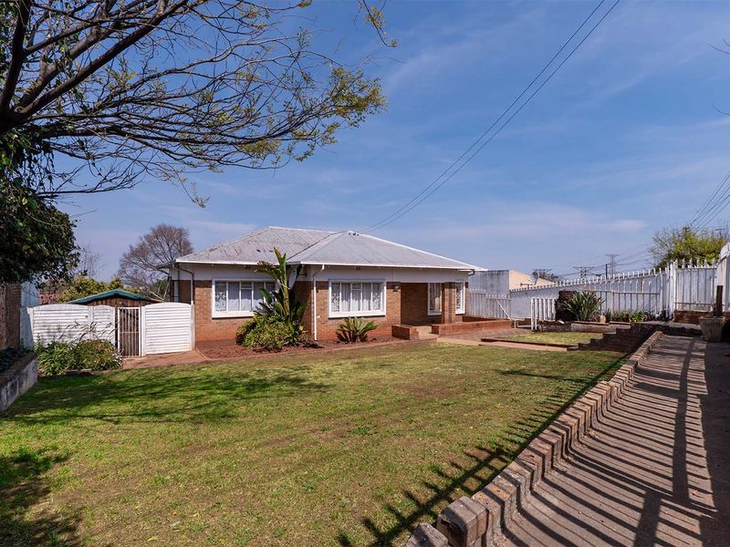 Front view of a single-story brick house featuring a corrugated metal roof, surrounding lawn, and perimeter fencing.