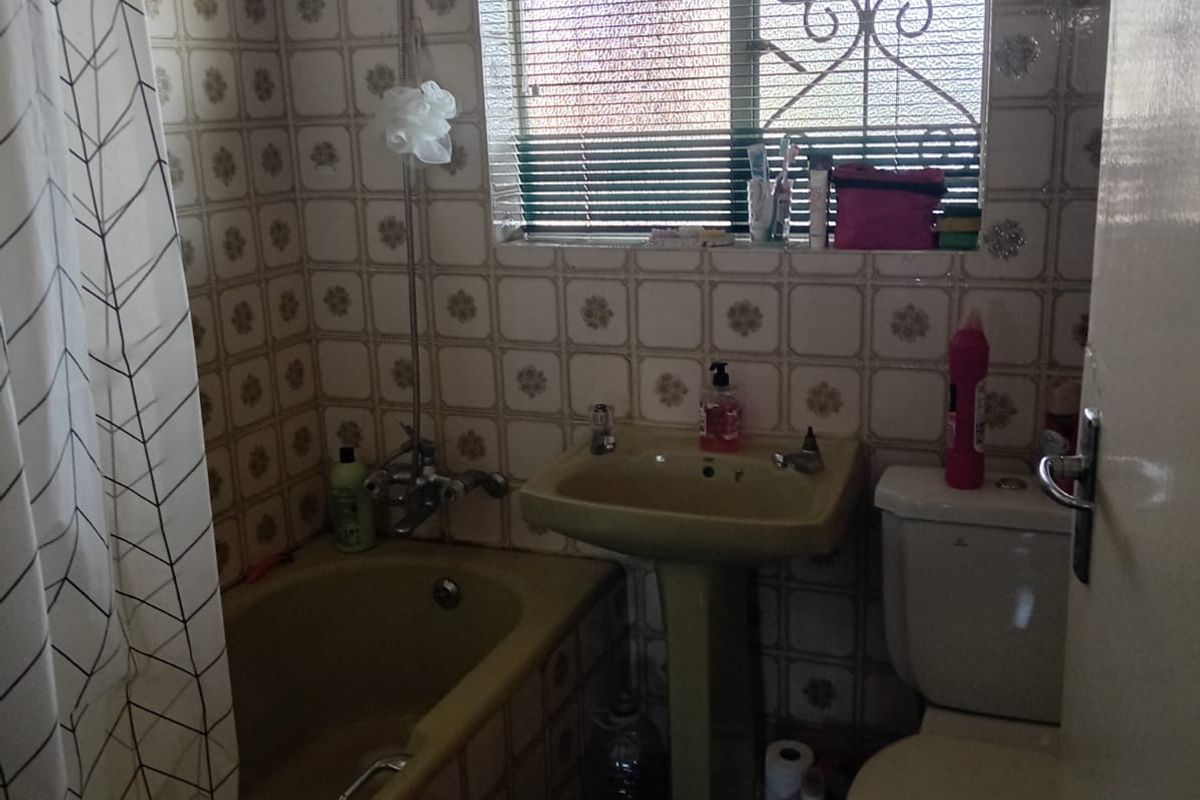 A compact bathroom featuring a pedestal basin, bathtub with shower mixer, and toilet, finished with dated wall tiling and a security-fitted window.