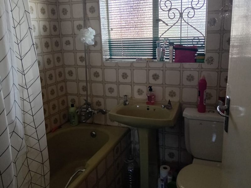 A compact bathroom featuring a pedestal basin, bathtub with shower mixer, and toilet, finished with dated wall tiling and a security-fitted window.