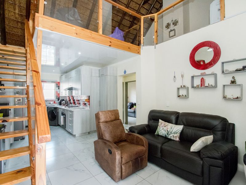 Open-plan living area featuring a modern kitchen, tiled floors, and a timber staircase leading to a mezzanine level with a thatched roof.