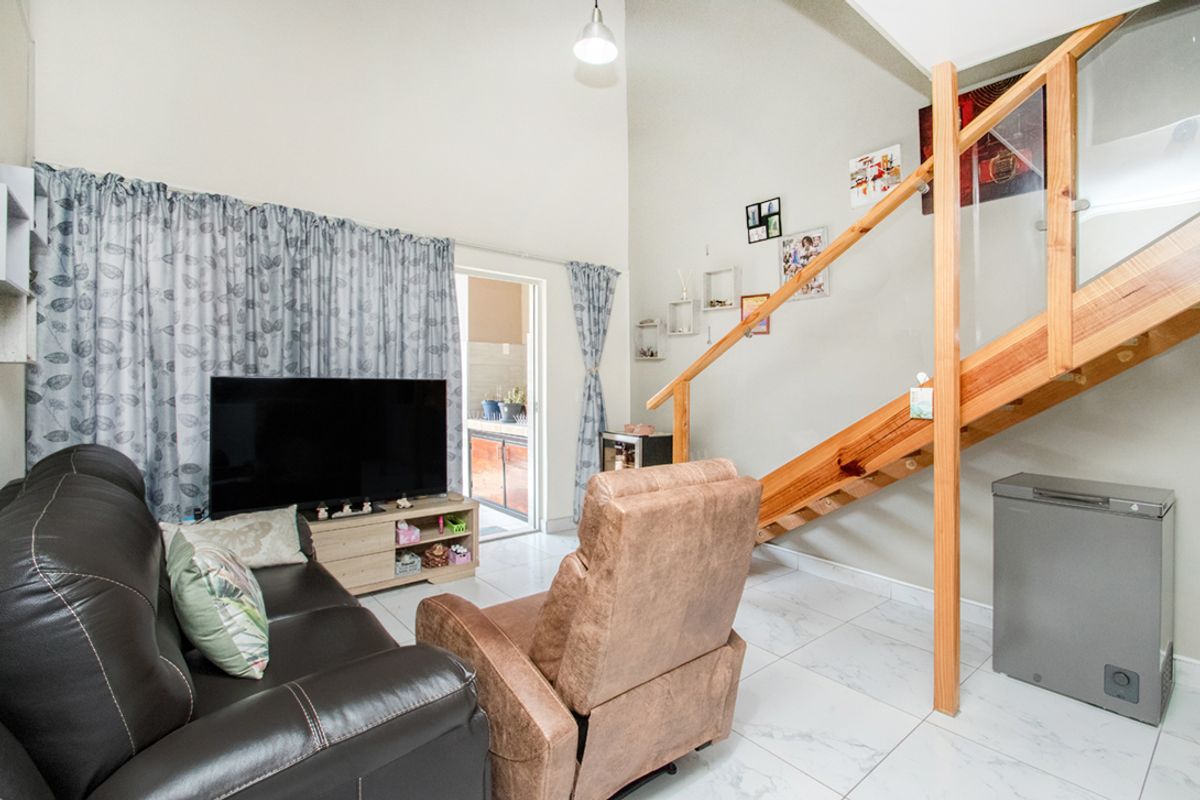 A bright living room featuring high vaulted ceilings, modern tiled flooring, and a wooden staircase with a glass balustrade leading to an upper level.