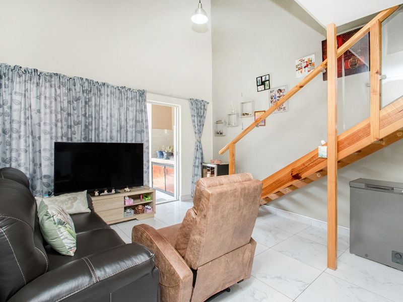 A bright living room featuring high vaulted ceilings, modern tiled flooring, and a wooden staircase with a glass balustrade leading to an upper level.