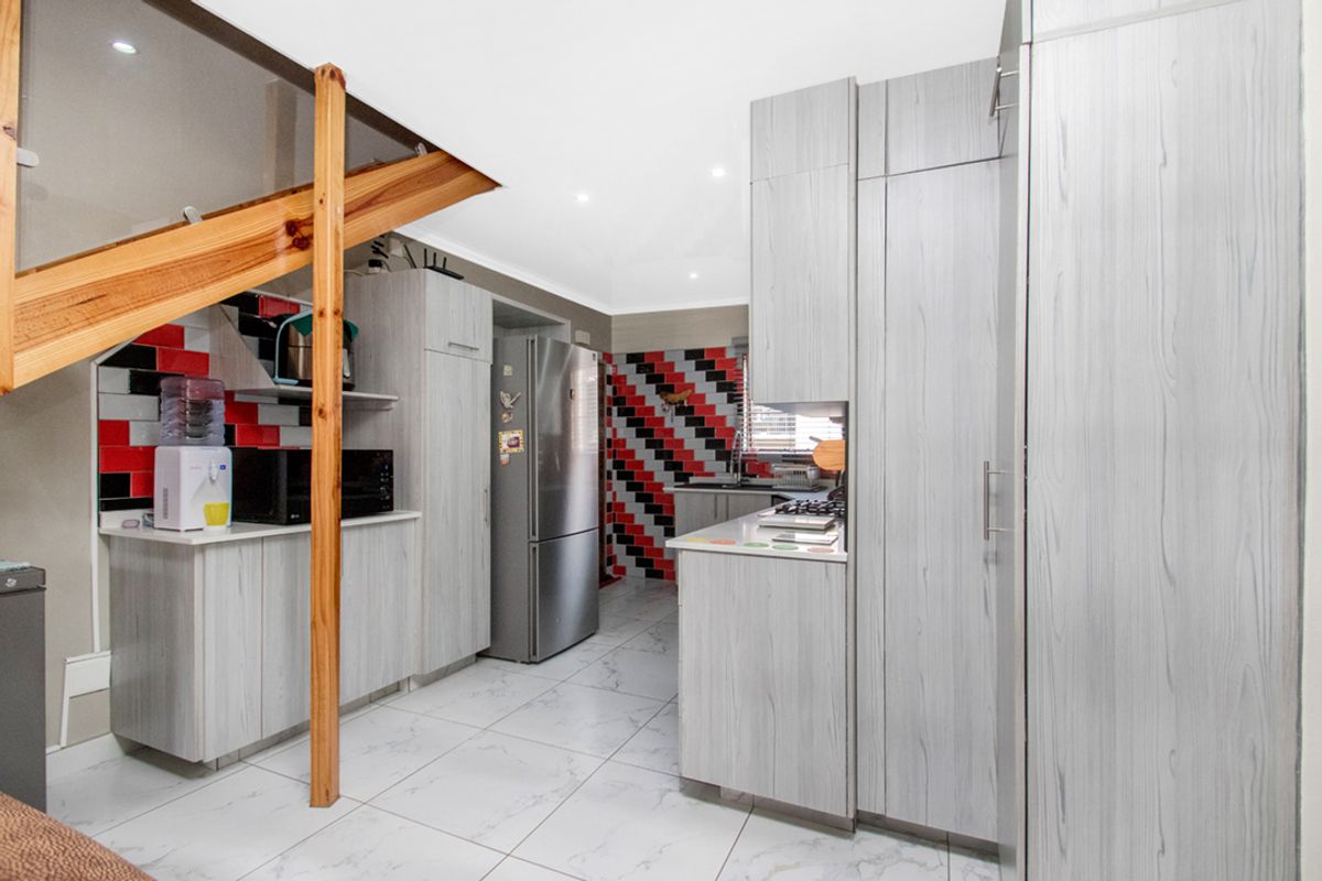 A functional kitchen featuring light wood-grain cabinetry, tiled flooring, and a distinctive red and black tiled backsplash, situated beneath a wooden staircase.