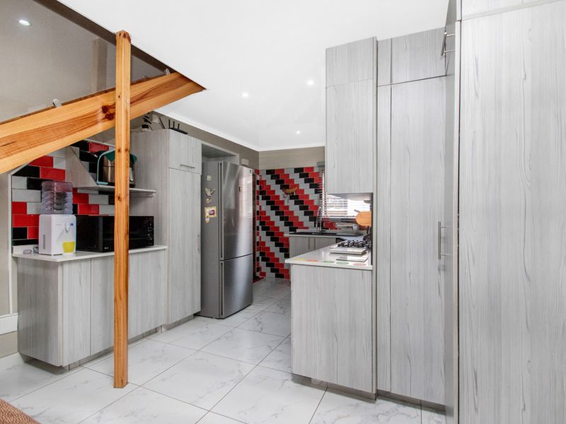 A functional kitchen featuring light wood-grain cabinetry, tiled flooring, and a distinctive red and black tiled backsplash, situated beneath a wooden staircase.
