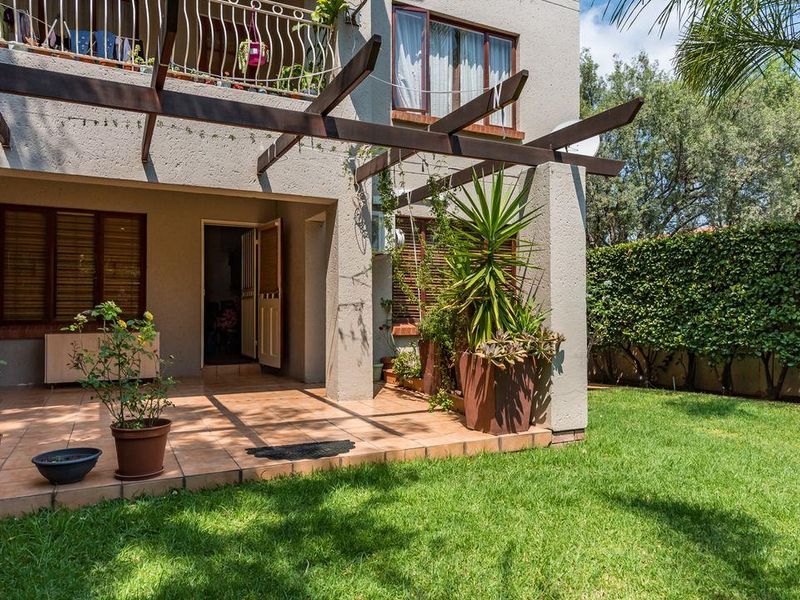A covered tiled patio area featuring a timber pergola, overlooking a private garden with established hedges and a balcony above.