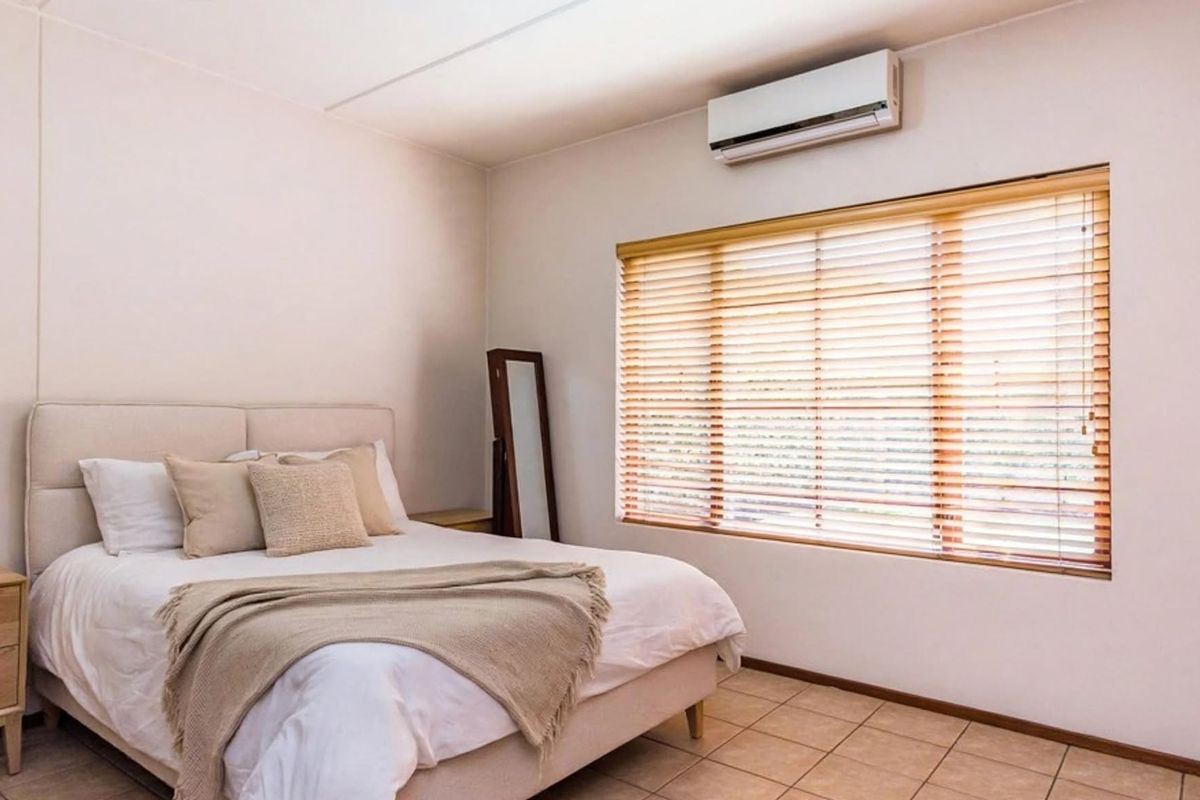 A well-lit bedroom featuring tiled flooring, a wall-mounted split-system air conditioner, and large windows fitted with wooden venetian blinds.