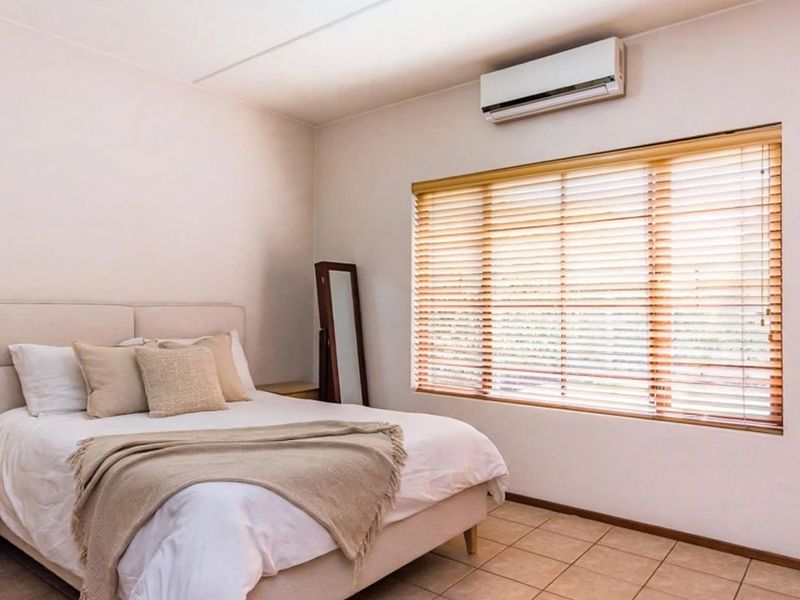 A well-lit bedroom featuring tiled flooring, a wall-mounted split-system air conditioner, and large windows fitted with wooden venetian blinds.