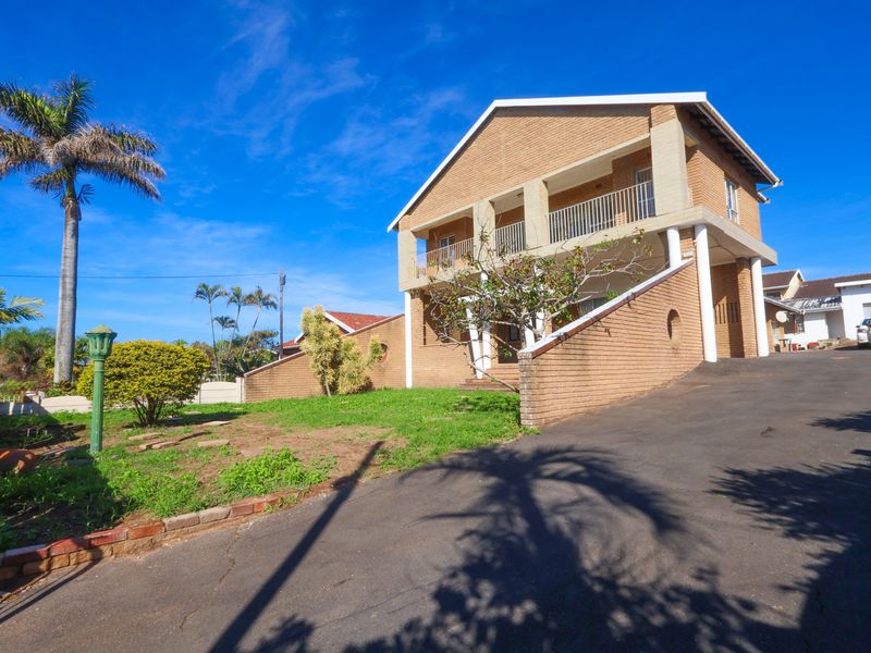 A double-story brick house featuring a front balcony, asphalt driveway, and established garden area with a retaining wall.