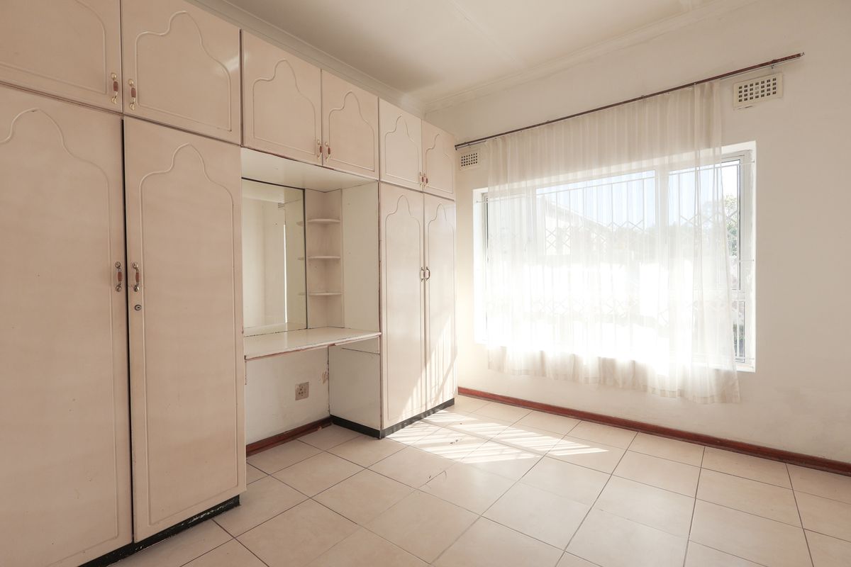 A bright, empty bedroom featuring extensive built-in cabinetry with an integrated vanity, tiled flooring, and a large window with security bars.