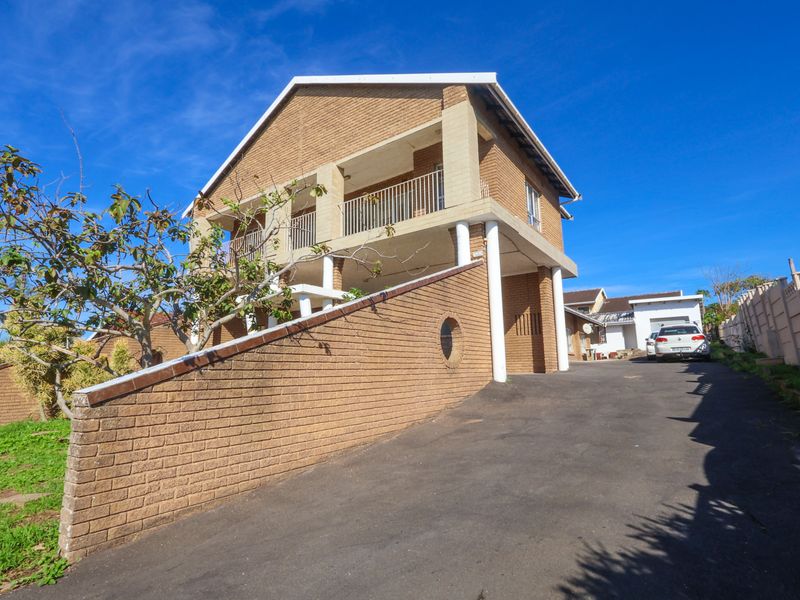 Front exterior view of a multi-level brick house featuring a paved driveway, elevated balcony with metal railings, and structural concrete pillars.