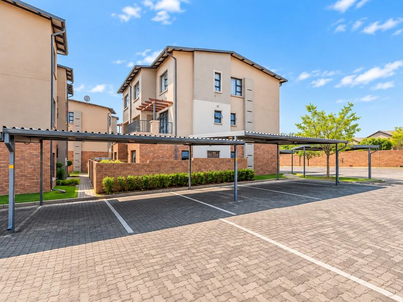 Exterior view of an apartment complex featuring covered parking bays, paved driveway, and multi-story residential buildings with brick and plaster finishes.