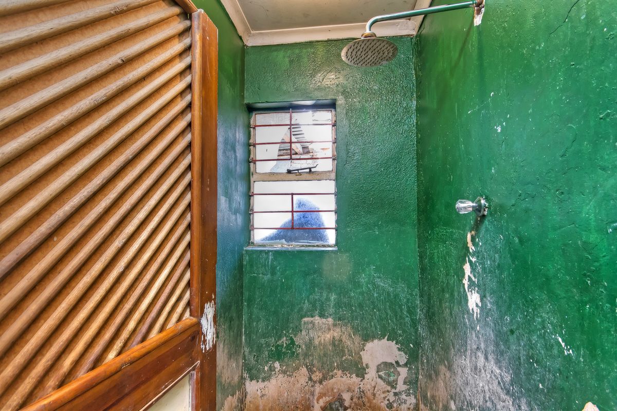 A small bathroom space featuring a shower head and a window with security bars, currently requiring significant wall repairs and cosmetic renovation.