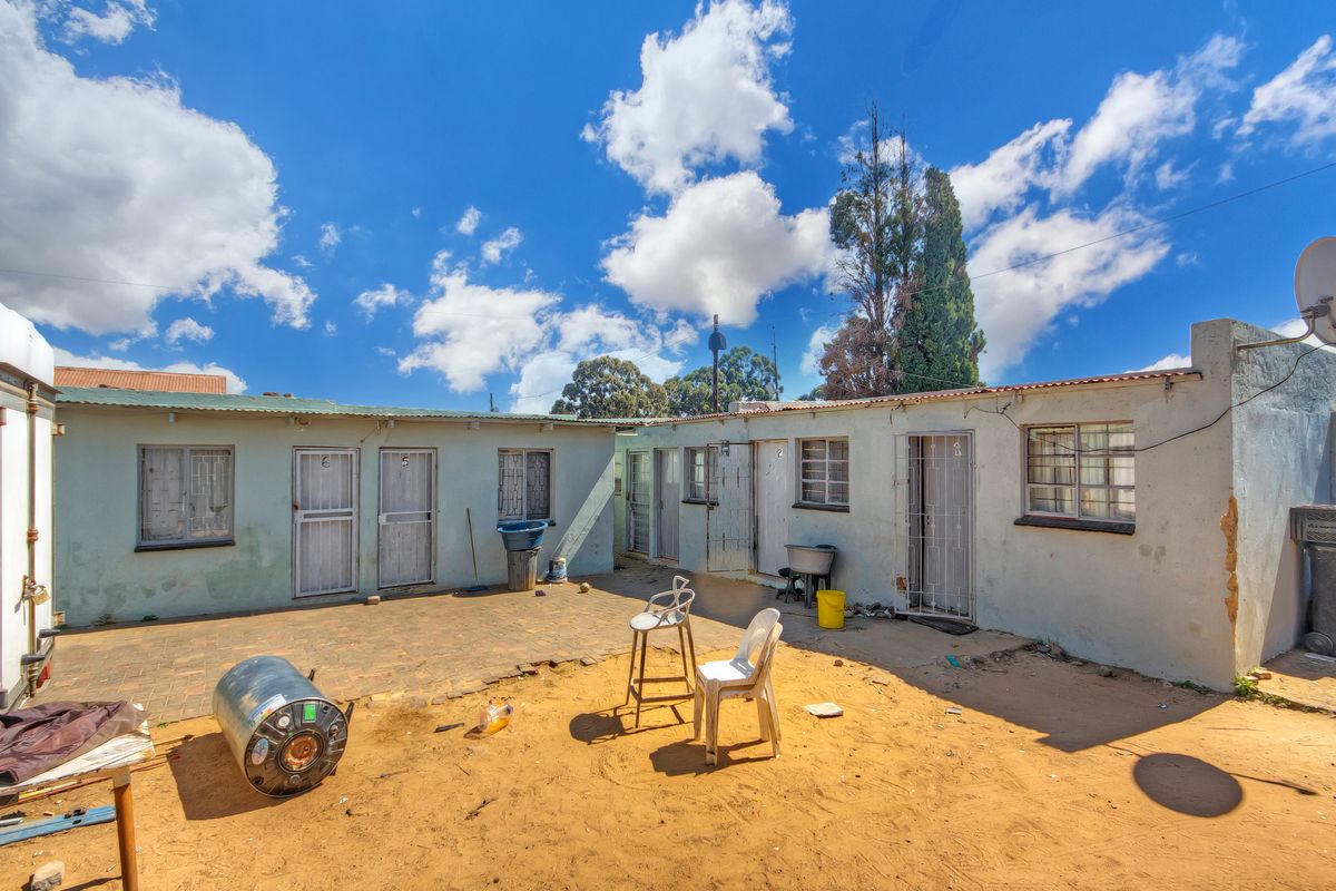 Rear exterior view of the property featuring a paved courtyard area, multiple access doors with security gates, and weathered exterior wall finishes.