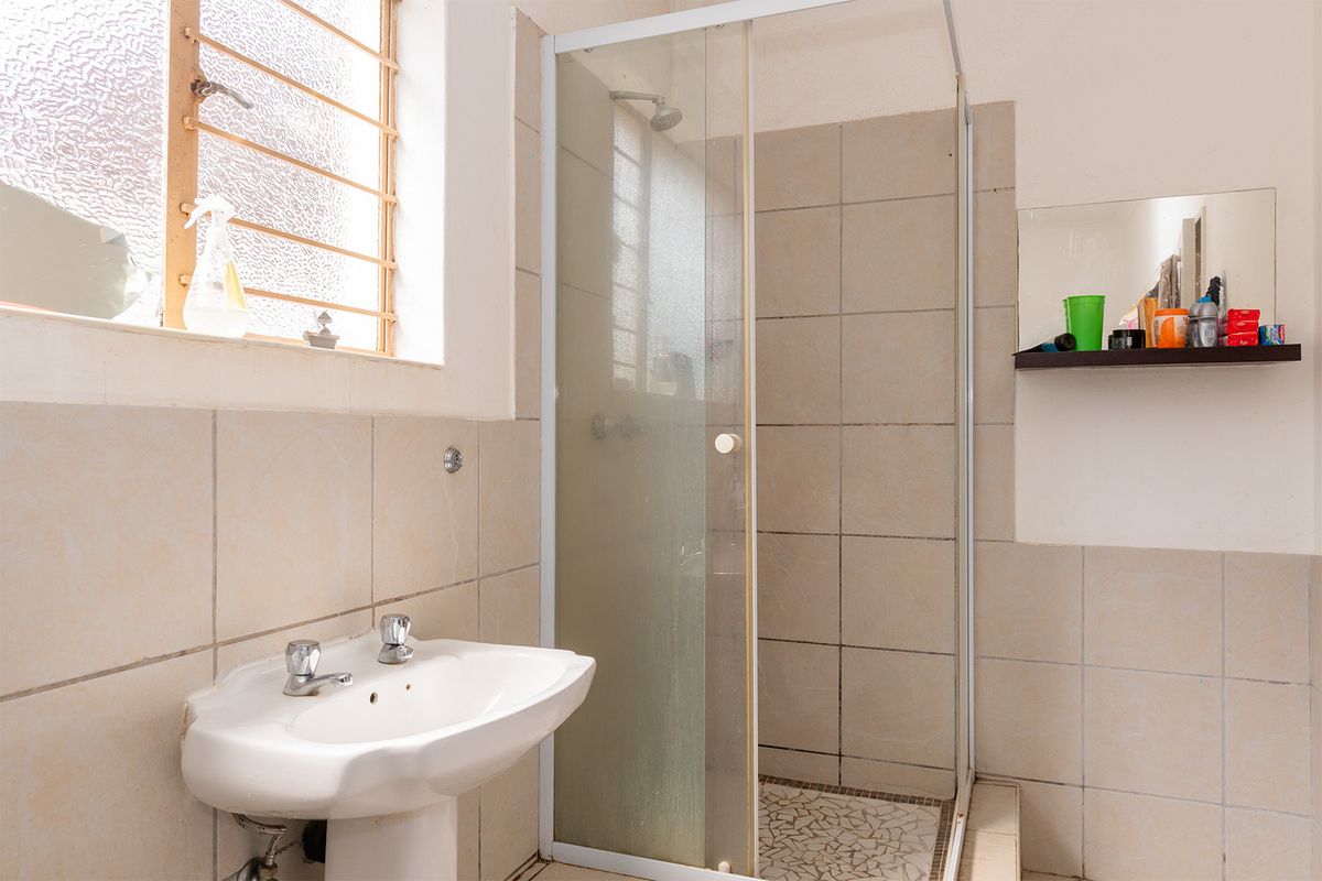 A functional bathroom featuring a pedestal washbasin, a glass-enclosed shower with tiled finishes, and a window with security bars.
