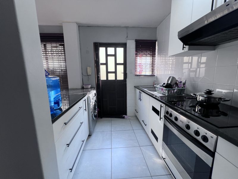 Functional galley-style kitchen featuring white cabinetry, granite countertops, tiled backsplash, and built-in electric stove with extractor fan, leading to an exterior door.