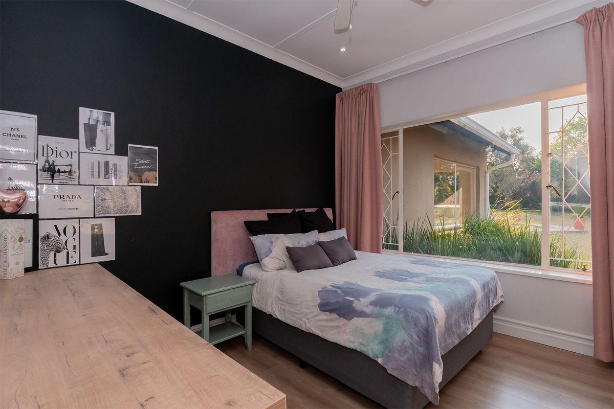 A well-lit bedroom featuring laminate flooring, a ceiling fan, and a large window with security bars overlooking the garden.