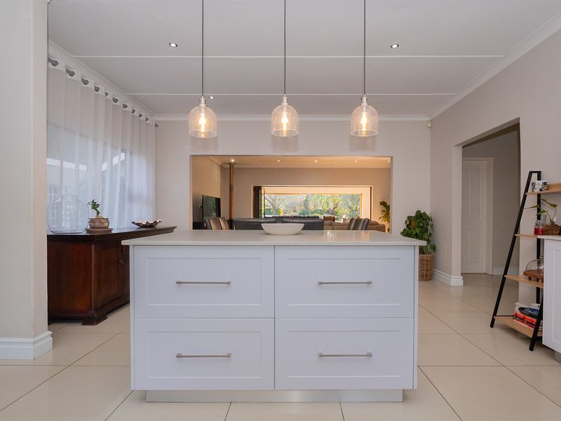 A modern, well-maintained kitchen featuring a central island with white cabinetry, sleek pendant lighting, and neutral tiled flooring, opening into the living area.