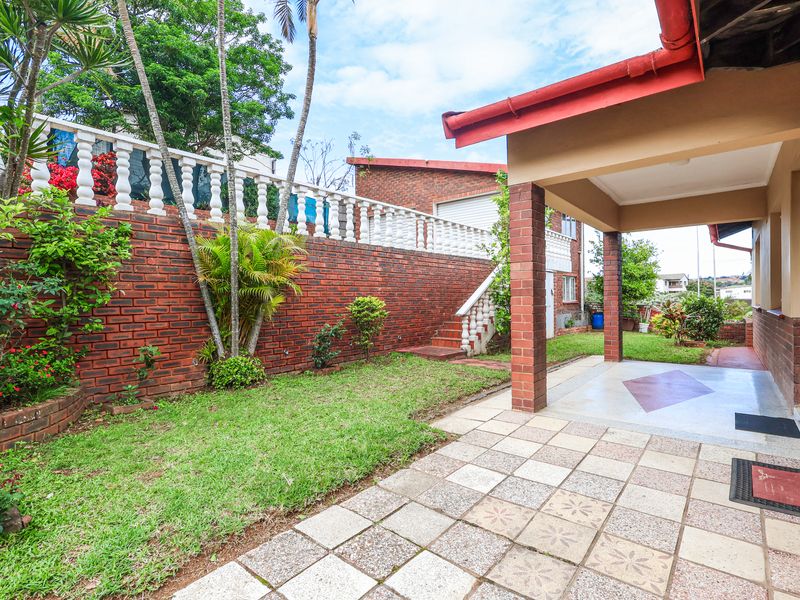 A covered patio area featuring tiled flooring and brick pillars, overlooking a landscaped garden with a raised brick retaining wall and balustrade.
