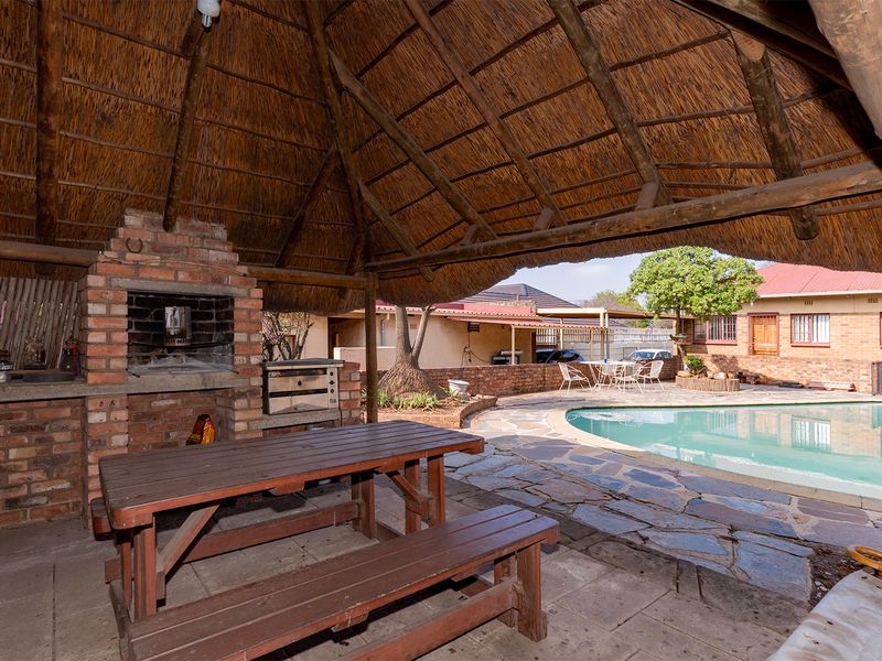 Covered outdoor entertainment area featuring a thatched roof, built-in brick braai, and direct access to a swimming pool and paved patio.