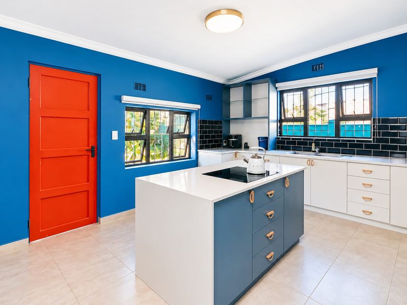 Modern kitchen featuring a central island with induction cooktop, white cabinetry, contrasting blue walls, and a bold red door.
