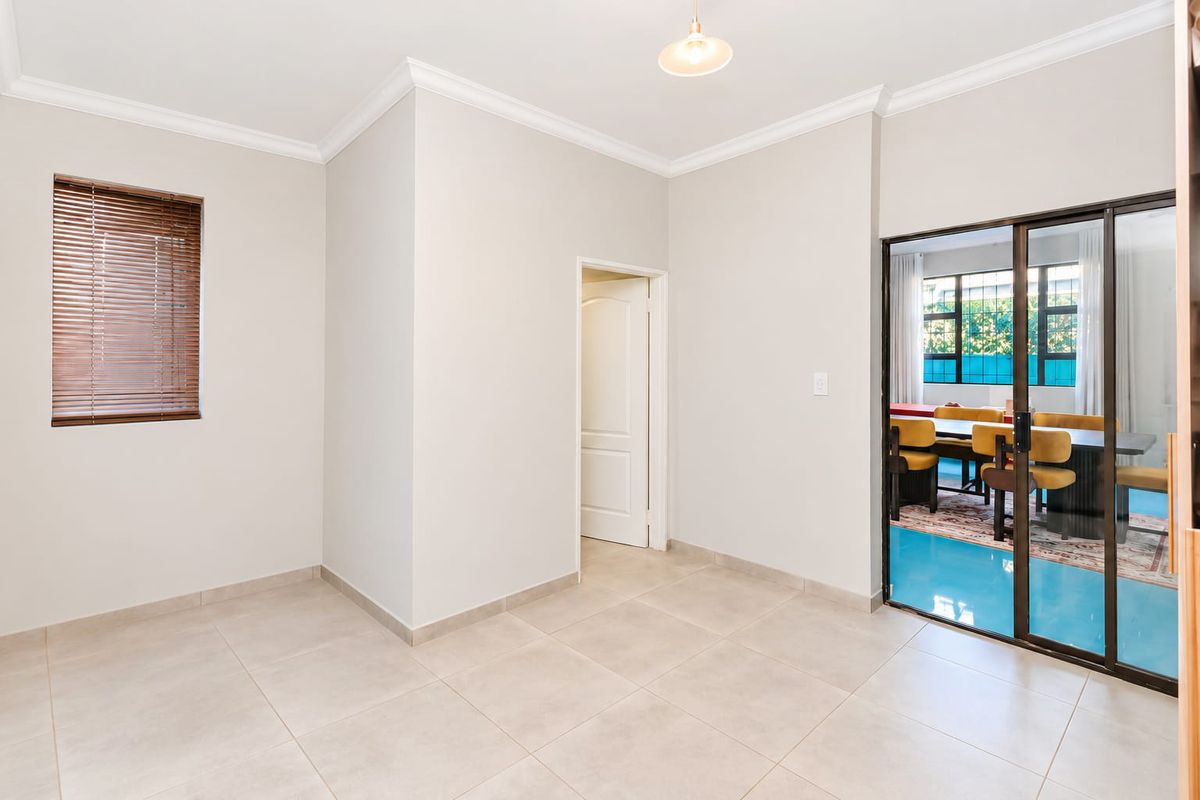A bright, empty living area featuring neutral tiled flooring, cove cornicing, and a sliding glass door providing access to an adjacent dining space.