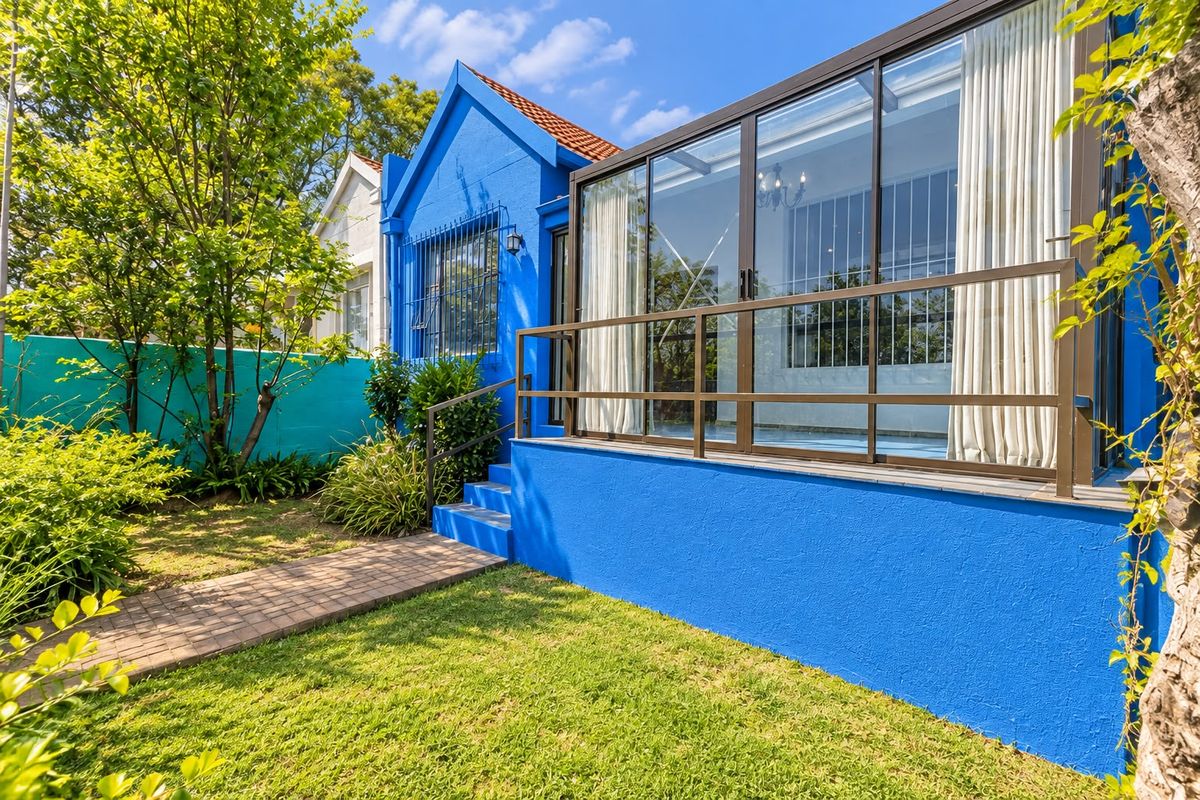 Rear view of the property featuring a vibrant blue exterior, a paved garden path, and an enclosed glass patio area with metal railings.