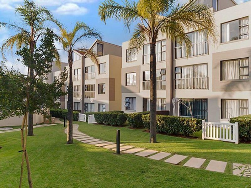 A well-maintained apartment complex exterior featuring landscaped gardens, paved walkways, and private balconies with sliding glass doors.
