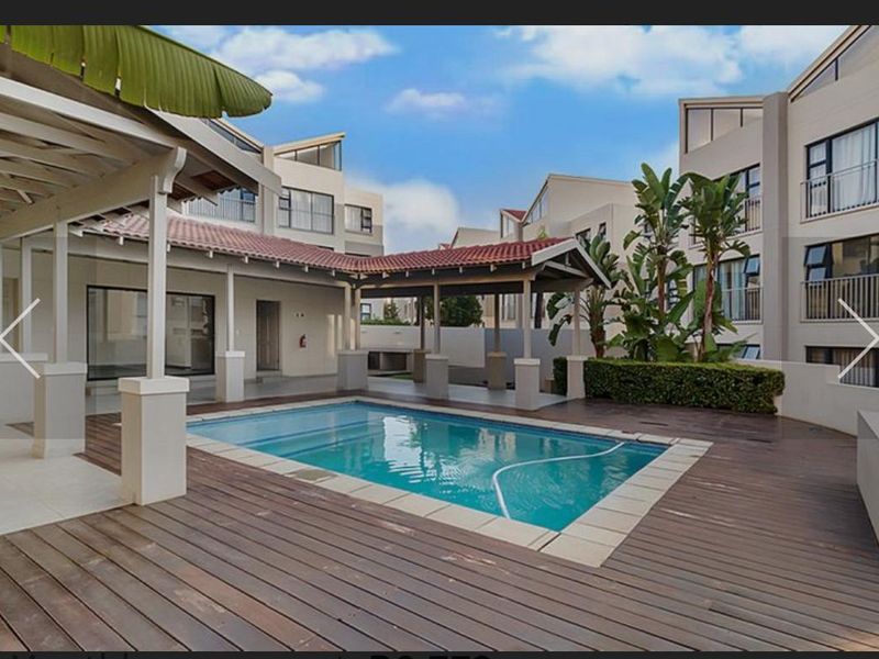 A well-maintained communal pool area featuring timber decking, a covered patio structure, and landscaped surroundings within an apartment complex.