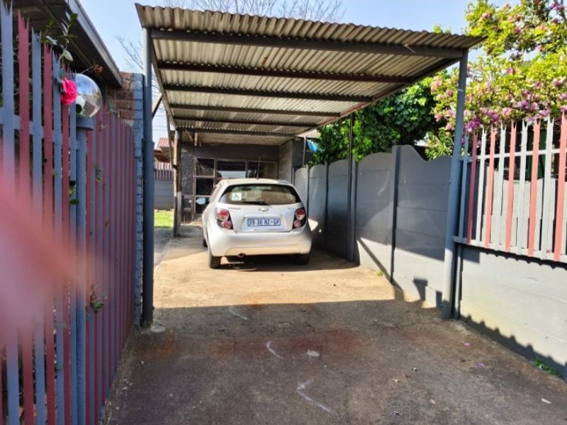 A covered carport area featuring a corrugated metal roof, concrete driveway, and perimeter security fencing.