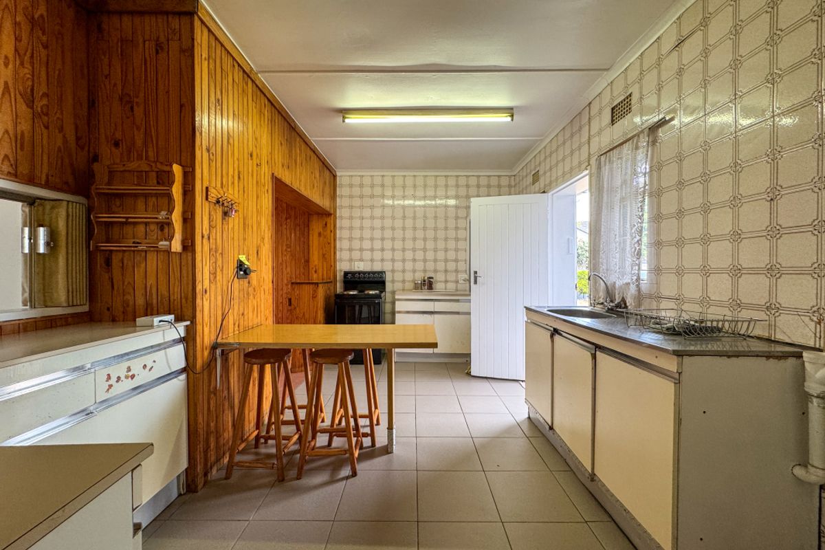 A dated kitchen featuring original timber wall paneling, tiled splashbacks, built-in cabinetry, and a functional layout with direct access to the exterior.