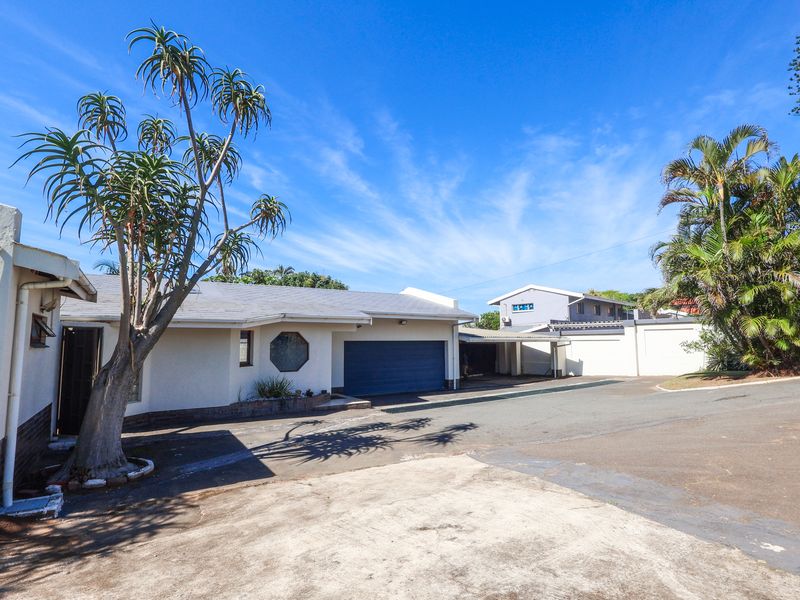 Front exterior view of the property featuring a paved driveway, attached garage, carport, and established garden area under a clear blue sky.