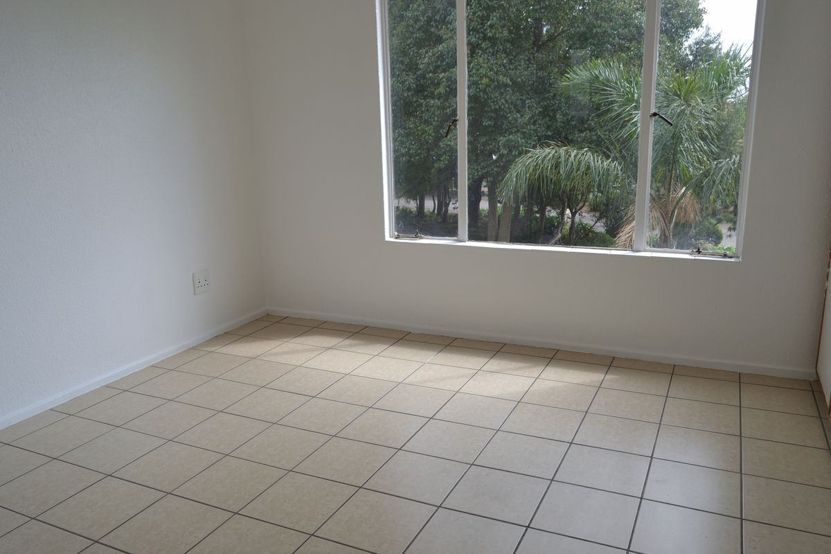 A bright, empty bedroom featuring neutral tiled flooring and a large window with a view of the surrounding greenery.
