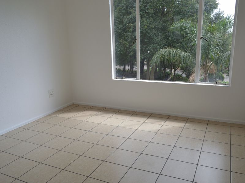 A bright, empty bedroom featuring neutral tiled flooring and a large window with a view of the surrounding greenery.