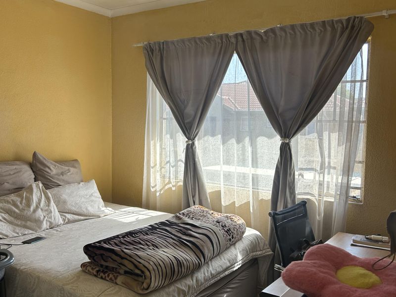 A bedroom featuring yellow walls, a window with curtains, and a ceiling light fixture. The room shows signs of wear on the ceiling and walls.