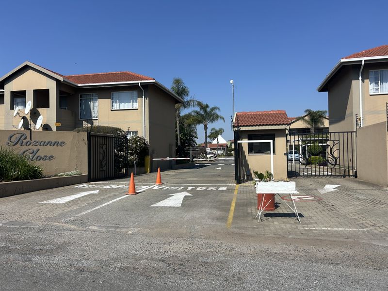 The entrance to Rozanne Close apartment complex featuring a secure gated access point, paved driveway, and established residential buildings.