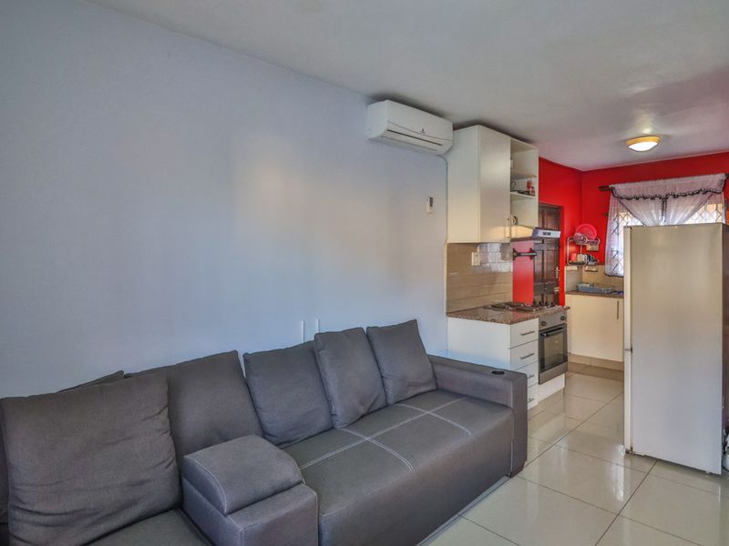 Open-plan living area featuring tiled flooring, a wall-mounted air conditioner, and a compact kitchen with built-in cabinetry and appliances.